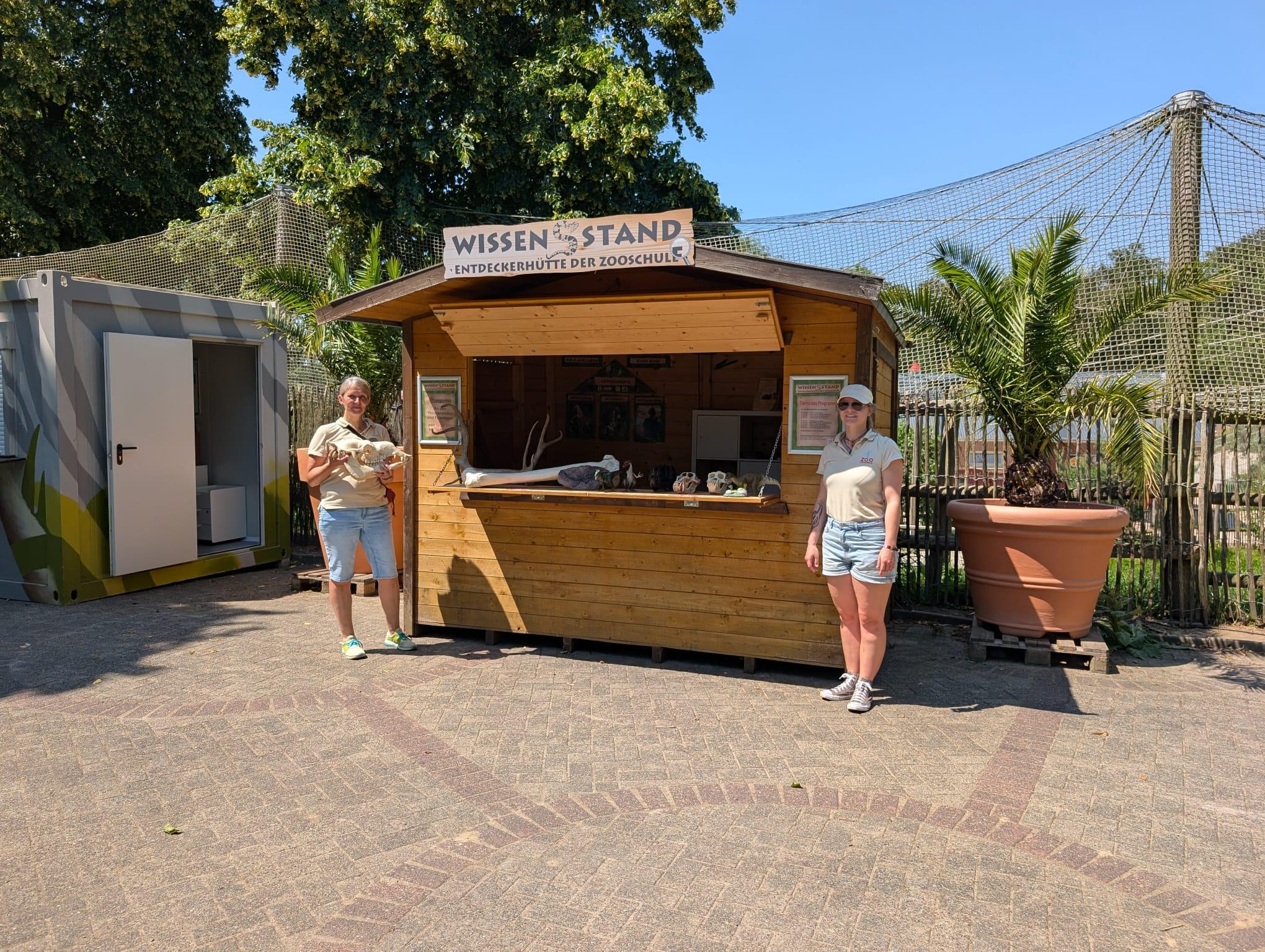 WissensStand am Pelikangehege Zweizoopädagoginnen stehen vor dem geöffneten WissensStand, einem Holzpavillon am Pelikangehege