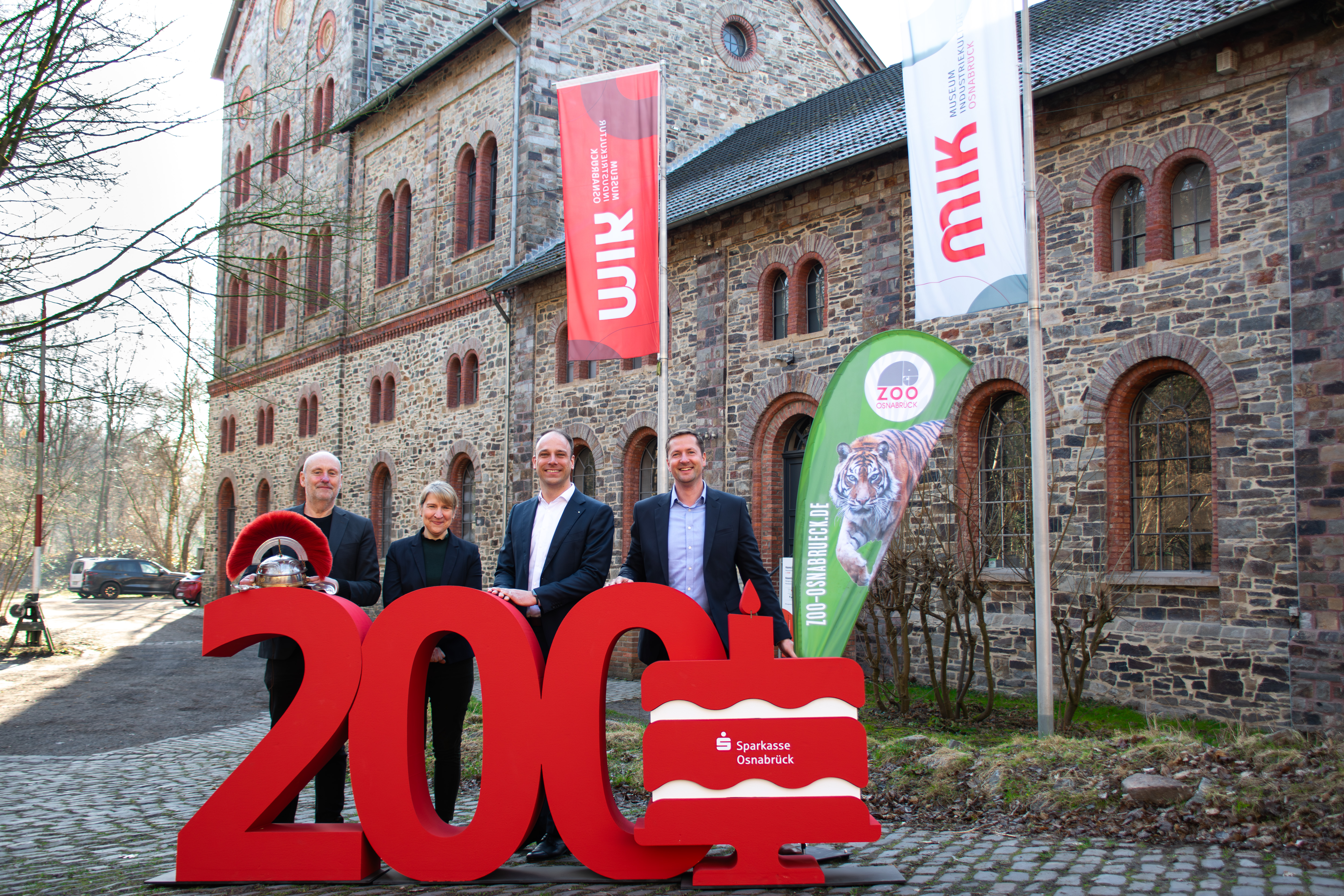 Vier Personen vor roter 200. Im Hintergrund sind drei Fahnen und ein Gebäude.