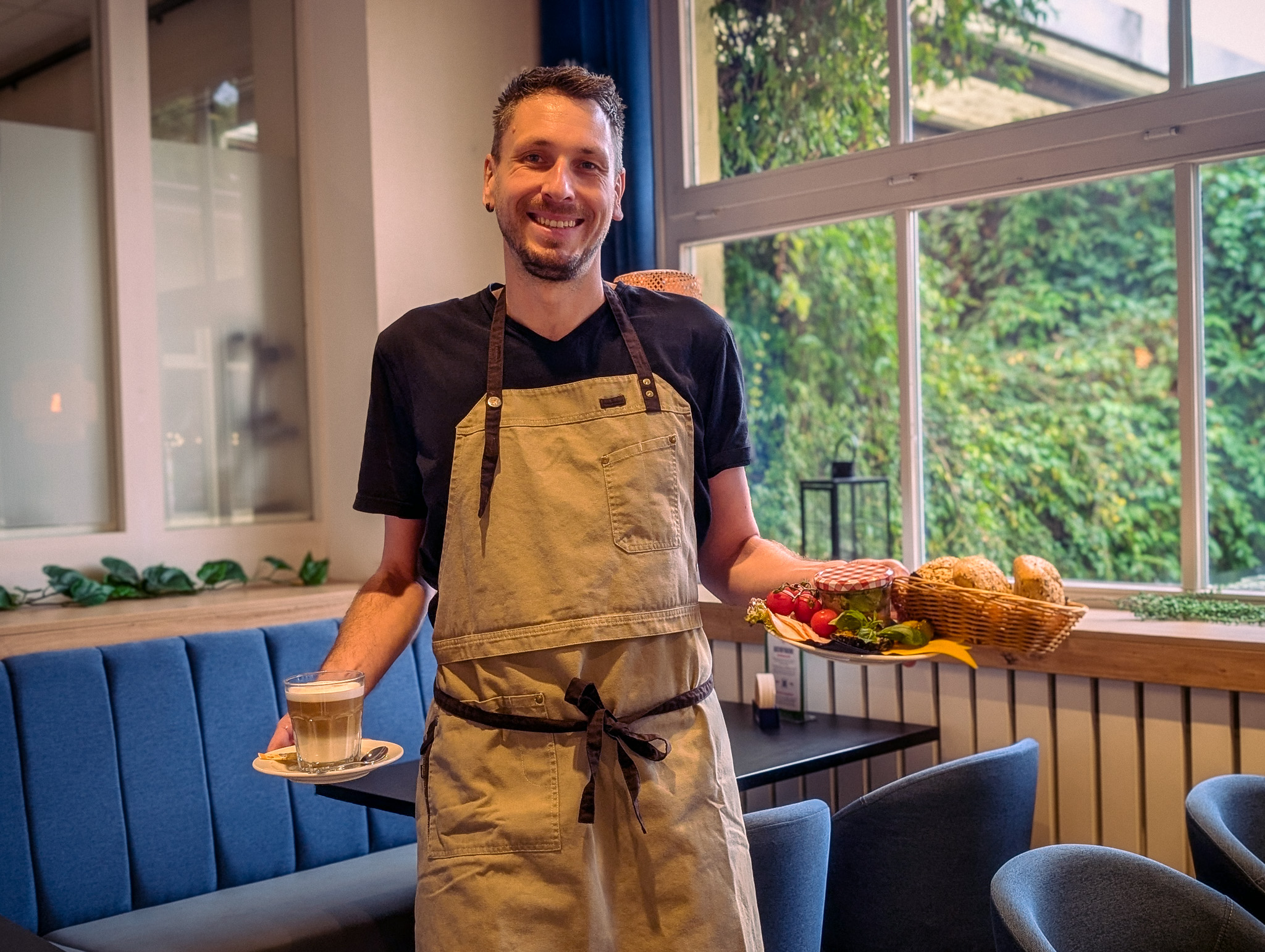 Sonntagsfrühstück im Zoo Ein Mann in einer Schürze gekleidet serviert ein Tablett mit Brötchen und frischen Frühstücksspeisen und einen Kaffee. Er lächelt herzlich in die Kamera.